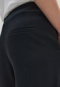 Svarta byxor i texturerat tyg med en enkel bakficka. Närbild på materialet och sömdetaljerna.