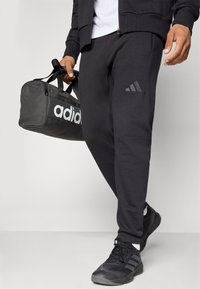 Férfi fekete Adidas melegítőnadrágot és sportcipőt visel, egy fekete Adidas tornazsákot tart a fogantyújánál egy sima háttér előtt.