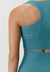 Turkusowy sportowy stanik o prążkowanej fakturze, z wycięciem i subtelnym logo z tyłu. Gładki materiał i dopasowany kształt.
