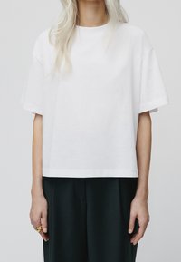 Camiseta de algodón blanca con un corte relajado, mangas cortas y dobladillo recto. La tela parece suave y ligera. Lleva pantalones oscuros.