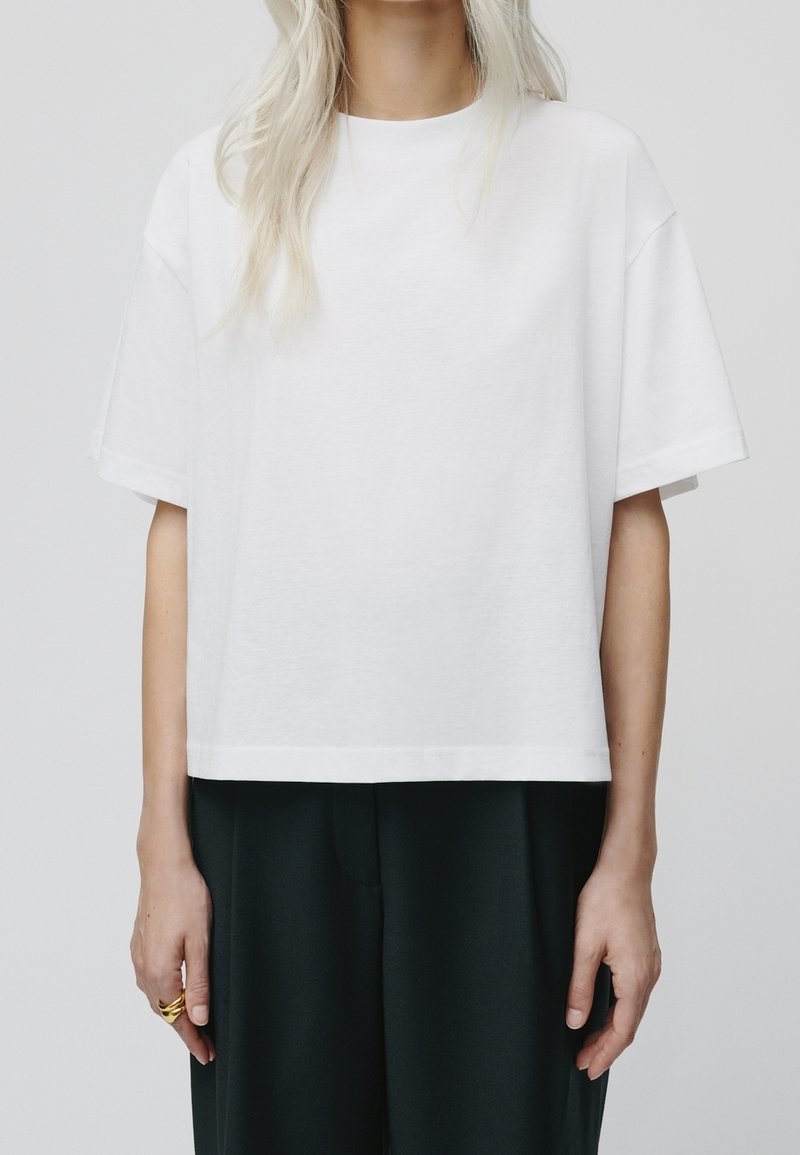 Camiseta de algodón blanca con un corte relajado, mangas cortas y dobladillo recto. La tela parece suave y ligera. Lleva pantalones oscuros.