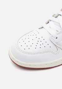 Adidași din piele albă cu cutie de degete perforată, șireturi albe plate și o talpă roșie contrastantă. Detalii cusute pe margine.