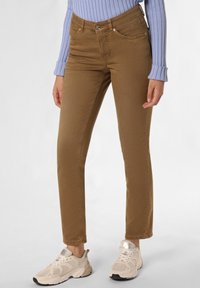 Jeans en denim marron avec une coupe ajustée, dotés de poches avant et d'une fermeture à bouton. Portés avec des baskets de sport beiges.