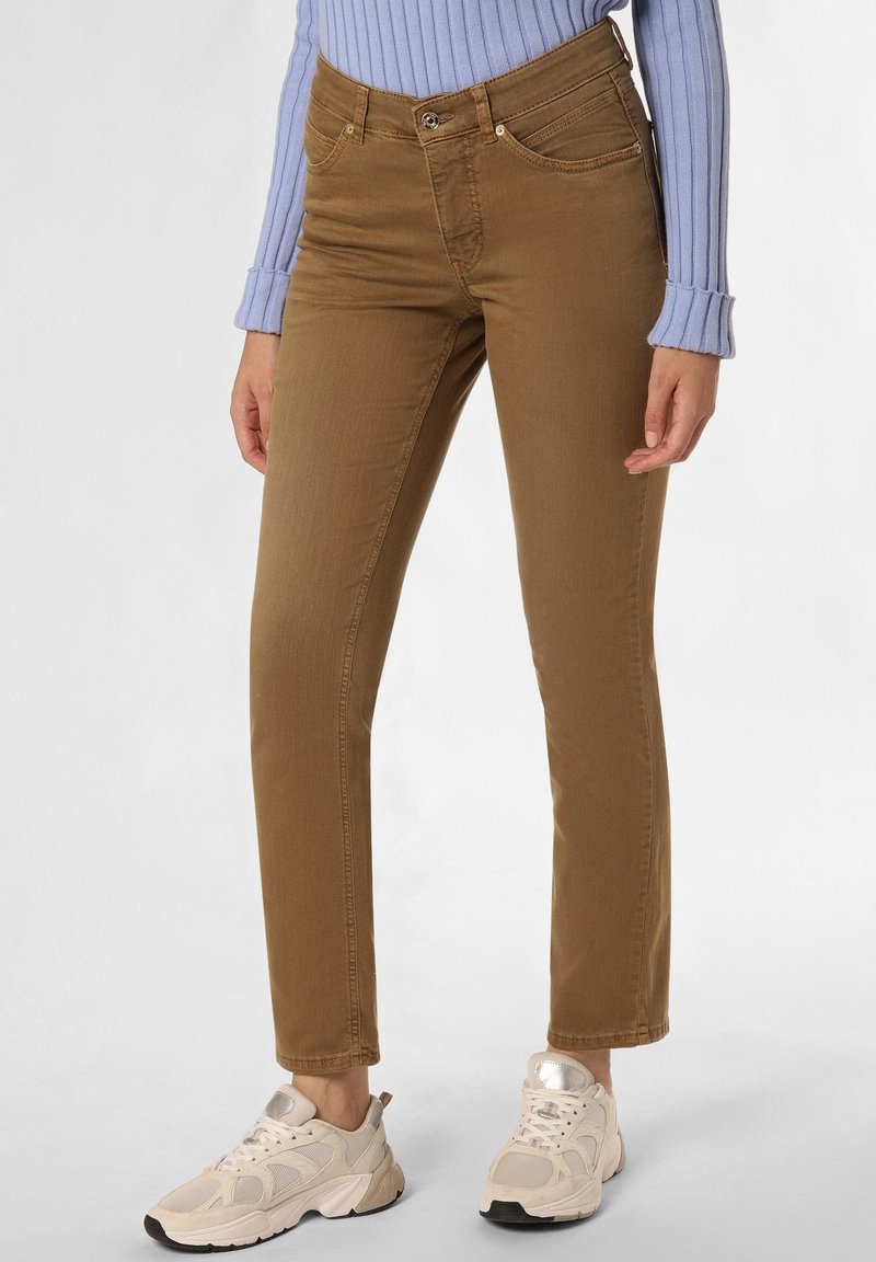 Jeans en denim marron avec une coupe ajustée, dotés de poches avant et d'une fermeture à bouton. Portés avec des baskets de sport beiges.