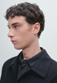 Cappotto nero con ampio colletto sovrapposto a una camicia grigia a bottoni, con motivo a righe sottili. Capelli corti e mossi; sfondo neutro.