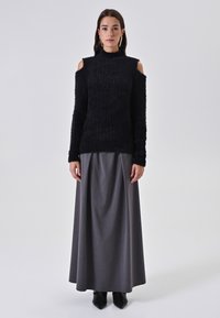 Maglione nero in maglia morbida con design a spalla scoperta, abbinato a una lunga gonna grigia. Il modello è in piedi di fronte a uno sfondo grigio chiaro.