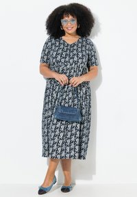Dunkelblaues, kurzärmliges Kleid mit weißem Paisleymuster; knielang. Kombiniert mit einer kleinen blauen Handtasche und blauen Ballerinas.
