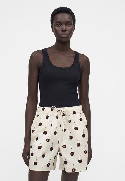 Γυναίκα φοράει μαύρο αμάνικο τοπ και κρεμ σορτς με κόκκινο σχέδιο κερασιών και κορδόνι στη μέση, στέκεται μπροστά σε απλό φόντο.