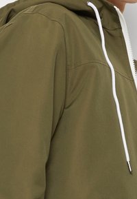 Chaqueta verde olivo con capucha, cordones blancos y cremallera en contraste. Presenta una textura suave y un detalle de costura minimalista.