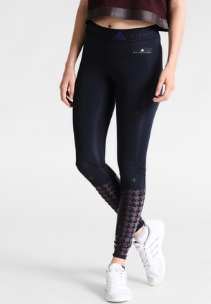 Person iført sorte atletiske leggings med mønstrede underben, hvide sneakers og en cropped mørkerød top mod en ensfarvet baggrund.