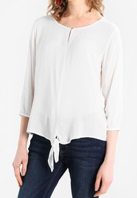 Blusa blanca con mangas tres cuartos, escote redondeado y un detalle de lazo en la parte frontal. Fabricada en un tejido ligero con una textura suave.
