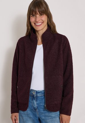 Femme souriante portant une veste texturée zippée violet foncé par-dessus une chemise blanche et un jean bleu sur fond uni.