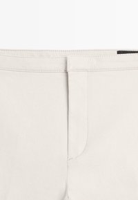 Taillenbereich einer hellbeigen Hose mit flacher Vorderseite und genähtem Bund, die ein minimalistisches, sauberes Design zeigt.