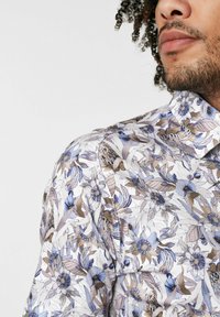 Hemd mit Blumenmuster, das blaue und braune Blumen auf einem weißen Hintergrund zeigt. Kurzer Kragen, knöpfbares Design und strukturierte Stoffdetails.