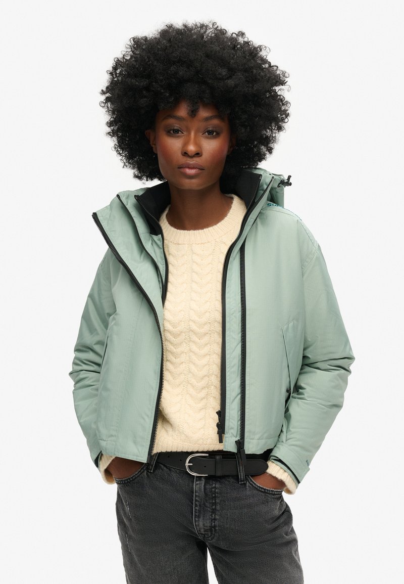 Lichtgroene jas met een rits aan de voorkant, verstelbare capuchon en zwarte accenten, gedragen over een crèmekleurige kabelbreiselsweater en grijze jeans.