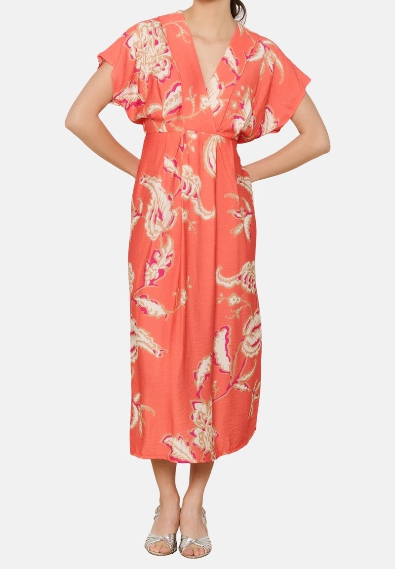 Femme portant une robe midi corail à imprimé fleuri, manches courtes, col en V et sandales argentées, debout les mains sur les hanches.
