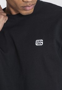 Camiseta de algodón negra con un logo bordado en blanco en el pecho. La tela tiene una textura suave y un corte clásico de cuello redondo.