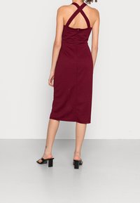 Robe midi bordeaux avec un motif texturé, col halter, bretelles croisées et silhouette ajustée, associée à des sandales à talons carrés noires.
