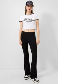 Witte cropped t-shirt met zwarte ringer en "Von Dutch Originals" graphic, gecombineerd met zwarte, aansluitende flare broek en zwart-witte sneakers.