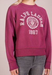 Person klädd i en magenta Ralph Lauren sweatshirt med "1967" och logotyptext, i kombination med blekta grå jeans, mot en enfärgad bakgrund.