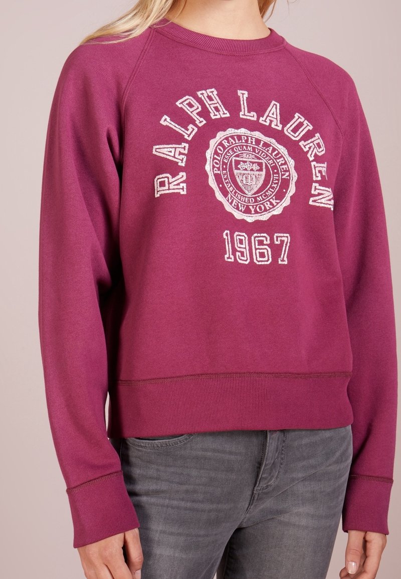 Person klädd i en magenta Ralph Lauren sweatshirt med "1967" och logotyptext, i kombination med blekta grå jeans, mot en enfärgad bakgrund.