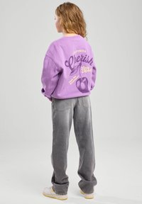 Violetti huppari, jossa on graafista tekstiä ja kuvastoa selässä, yhdistettynä haalistuviin harmaisiin farkkuihin ja valkoisiin sekä keltaisiin lenkkareihin.