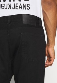 Černé džíny vyrobené z denimové látky, s koženou nášivkou na pase, zadními kapsami a rovný střihem. Textura se zdá být hladká s detaily šití.