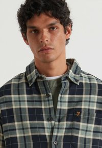 Camisa de botones a cuadros en tonos azul oscuro y verde, con un tejido texturizado, cuello tradicional y un logo bordado en el pecho.
