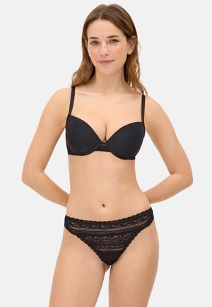 Mujer modelando un sujetador negro acolchado con tirantes finos y braguitas de encaje a juego sobre un fondo claro liso.