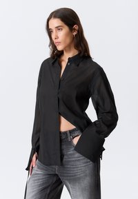 Chemise noire boutonnée en tissu léger, avec une coupe ample, des manches longues et des fentes latérales pour plus de détails. Associée à un jean gris.