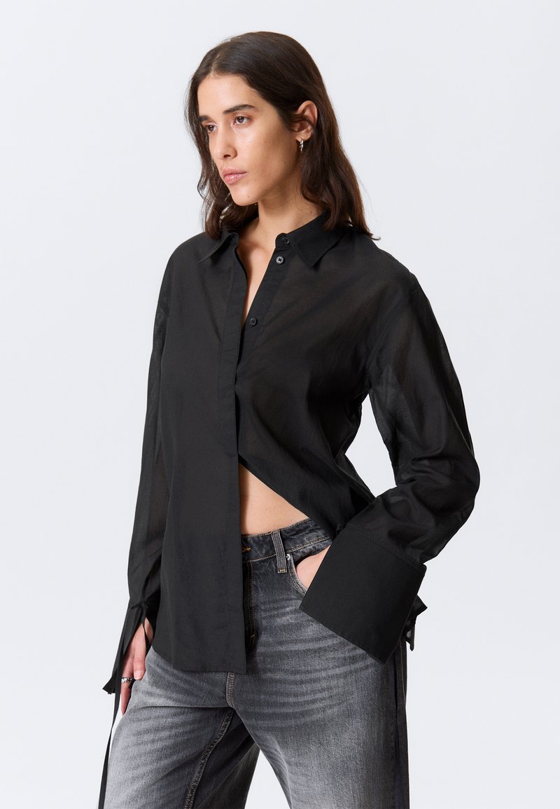 Chemise noire boutonnée en tissu léger, avec une coupe ample, des manches longues et des fentes latérales pour plus de détails. Associée à un jean gris.