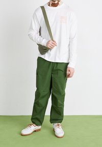 Vit långärmad t-shirt med grafiskt tryck, gröna cargobyxor med fickor, vita skor med bruna detaljer, och en olivgrön axelväska.