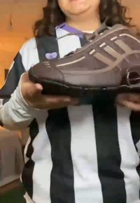 Brązowy but sportowy o zakrzywionym kształcie, fakturowanej powierzchni i trzema białymi paskami. Trzymany przez osobę ubraną w czarno-białą koszulę w paski.