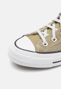 Plátěné tenisky v khaki barvě s bílou gumovou špičkou a podrážkou, šedými šňůrkami a černým lemem podél podrážky. Obsahují prošívané detaily.