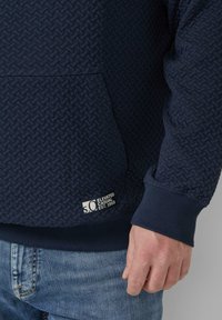 Felpa blu navy con trama a rete, polsini a costine e tasca frontale. Presenta un'etichetta con la scritta "S.C. Elevated Casual Est. 1969."