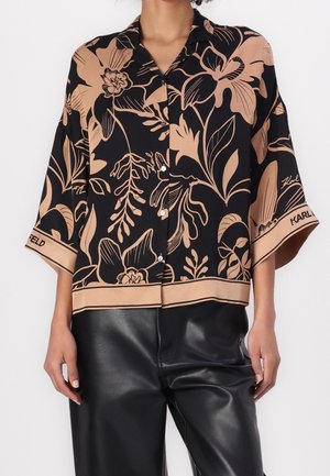 Kobieta ubrana w czarno-beżową kwiecistą bluzkę na guziki z szerokimi rękawami oraz czarne skórzane spodnie, stojąca na białym tle.
