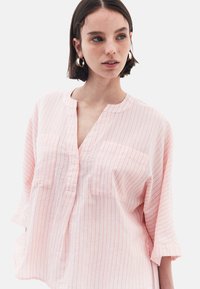 Camicia leggera a righe in rosa chiaro e bianco. Presenta un collo a V, due tasche sul petto e maniche 3/4. Realizzata in materiale traspirante.
