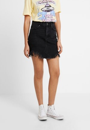 Personne vêtue d’un t-shirt graphique jaune, d’une mini-jupe en denim noire effilochée et de baskets blanches montantes, debout devant un fond uni.