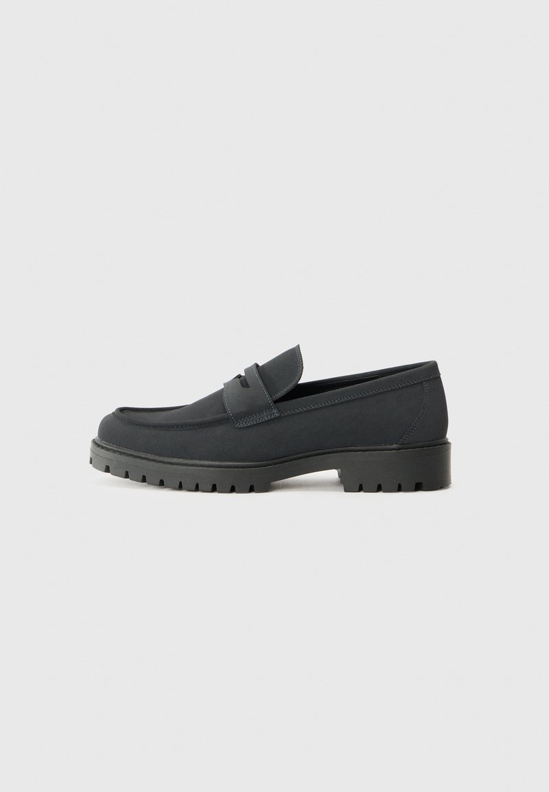 Mocassin en cuir noir avec une semelle épaisse à crampons et un détail de lanière sur le dessus, présenté de côté extérieur sur un fond uni.