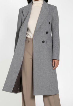 Kobieta ubrana w długi szary płaszcz dwurzędowy na kremowym golfie i beżowych spodniach na białym tle.