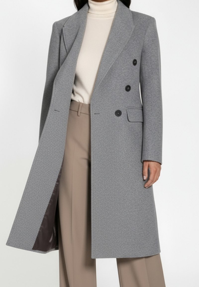 Femme portant un long manteau gris croisé sur un col roulé crème et un pantalon beige sur fond blanc.