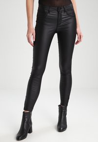 Jean skinny noir en matériau brillant et élastique, doté de cinq poches, associé à des bottines noires texturées.