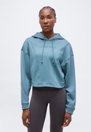 Kvinde med fletninger iført en blå Nike Pro hættetrøje og mørkegrå leggings, stående foran en ensfarvet hvid baggrund.