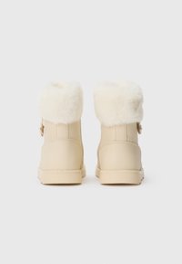 Bottes d'hiver beiges avec des poignets en fausse fourrure blanche, semelles robustes et coutures visibles, vues de dos sur un fond uni.
