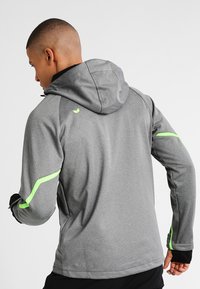 Veste de sport grise avec capuche, tissu texturé et accents vert fluo sur les côtés et les manches. Poignets ajustables inclus.