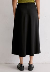 Schwarzer Midi-Rock aus glattem, fließendem Stoff mit einem Reißverschluss hinten. Getragen mit schwarzen Schuhen, die eine flache Sohle und ein minimalistisches Design aufweisen.
