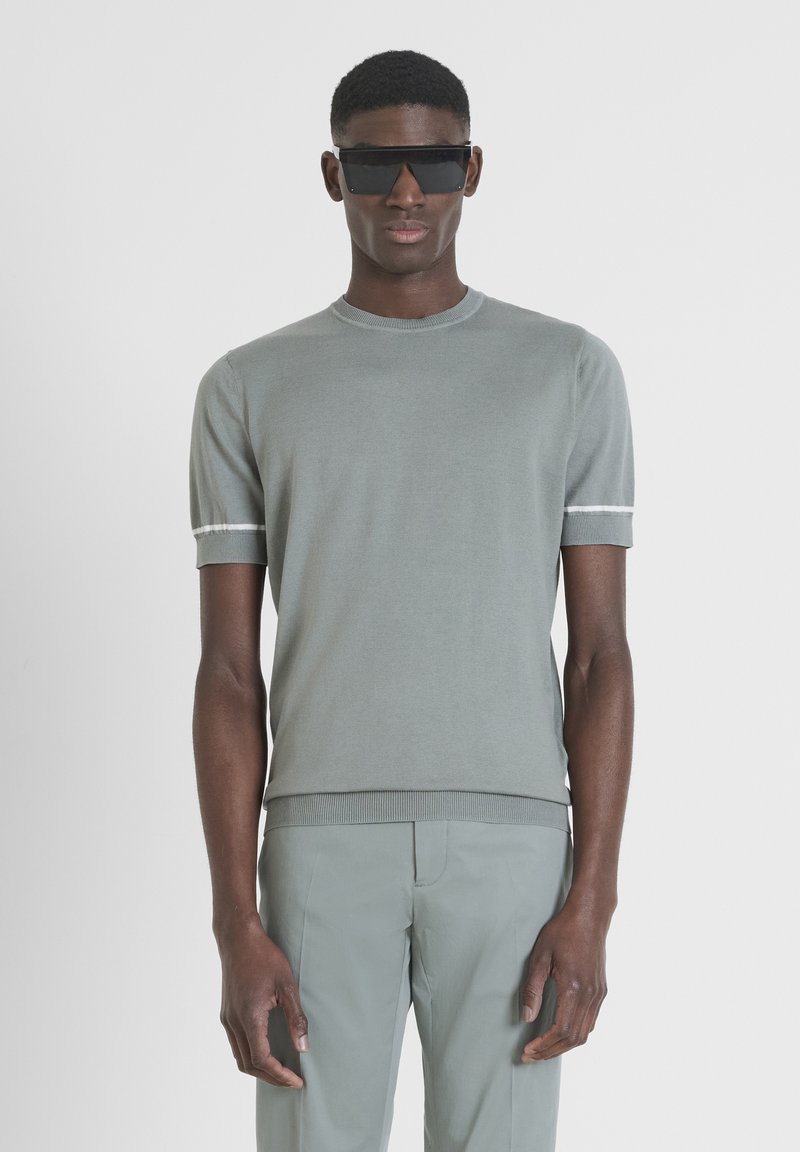 Maglione a maglia grigio chiaro con maniche corte, caratterizzato da strisce bianche sui polsini e un collo rotondo. Indossato con pantaloni grigio chiaro abbinati.