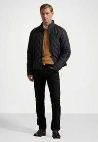 Μαύρο καπιτονέ μπουφάν με όρθιο γιακά, σε συνδυασμό με πουλόβερ μουσταρδί με πλέξη καλωδίου, μαύρο τζιν και μαύρα παπούτσια, παρουσιάζεται σε ουδέτερο φόντο.