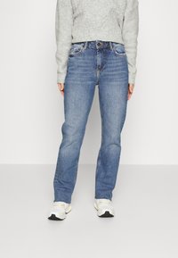 Jeans de ganga azul com corte reto, cinco bolsos e desbotamento subtil. Combinados com um sweatshirt texturizado cinza claro e ténis brancos.