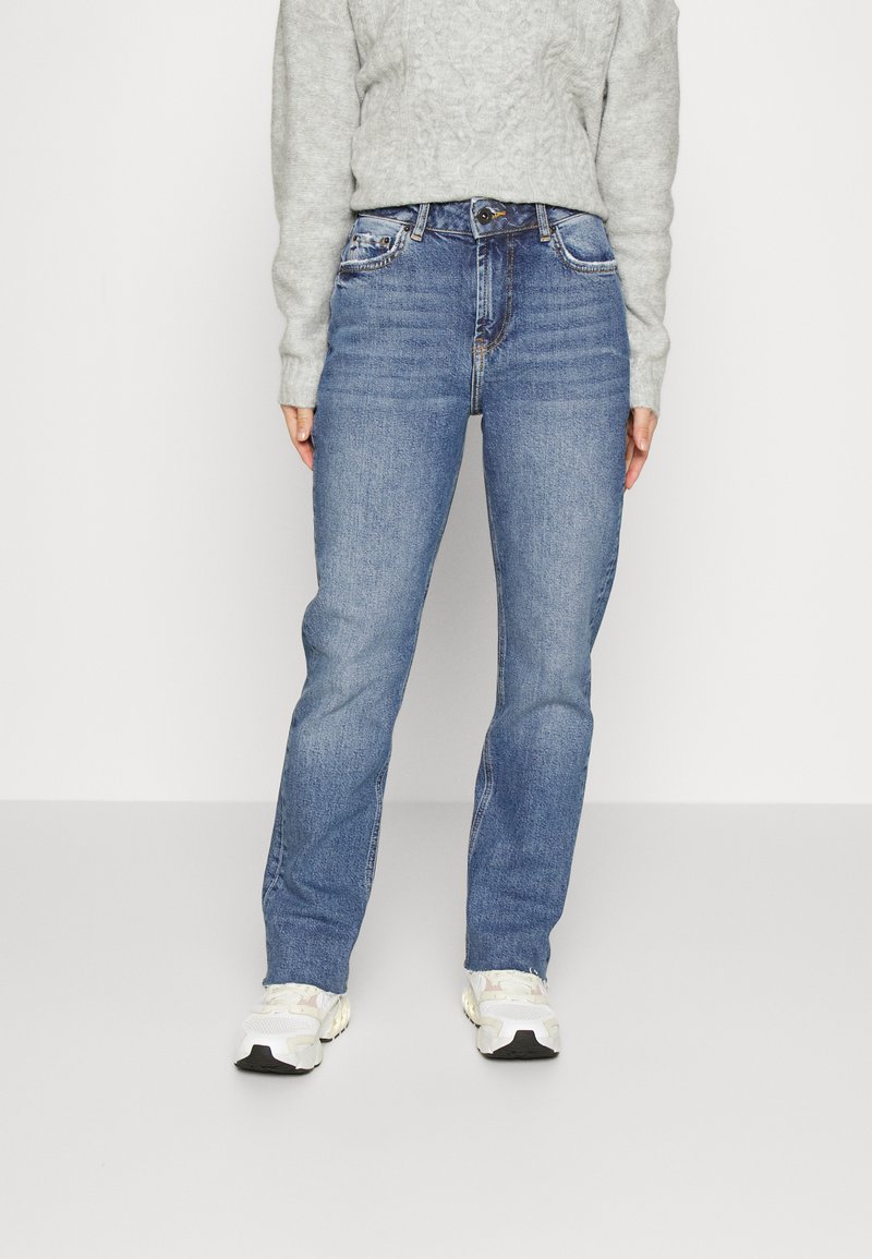 Jeans de ganga azul com corte reto, cinco bolsos e desbotamento subtil. Combinados com um sweatshirt texturizado cinza claro e ténis brancos.